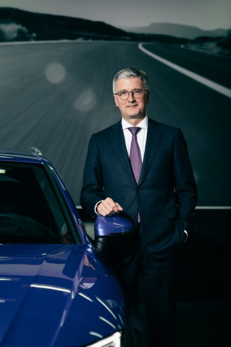 Rupert Stadler mit Hand auf dem Außenspiegel eines blauen Audi A4 Quattro vor dem Bilder einer Bergstraße.